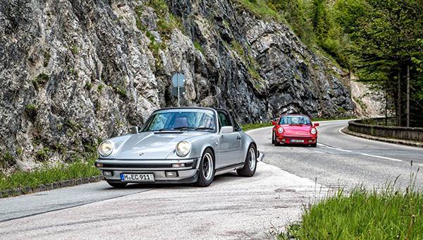 Zwei Porsche 911 fahren hintereinander auf Straße neben einer Felswand