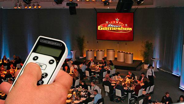 Hand with remote control pointing at view of restaurant with people having dinner, Dinner Gameshow logo on a screen in the background