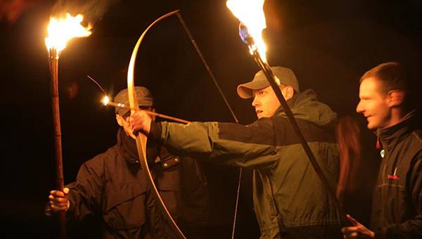 Two people explaining to one man about archery holding a crossbow and an arrow with flaming tip
