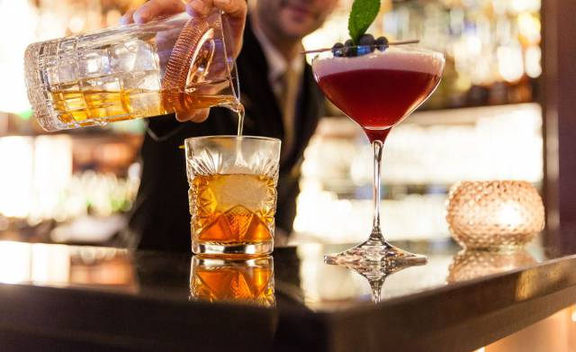 Two cocktails on a bar at night time, one is being topped up by bartender