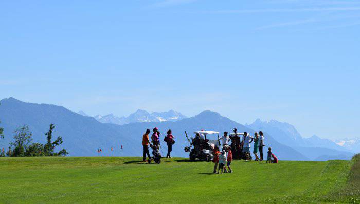 Gruppe Golfer mit Kart vor Loch mit Alpenpanorama